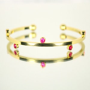Peut inclure: Un bracelet jonc doré orné de petites pierres précieuses roses rondes, espacées régulièrement sur le bracelet. Le bracelet se reflète sur une surface brillante, créant un effet miroir. Bijou simple et élégant.