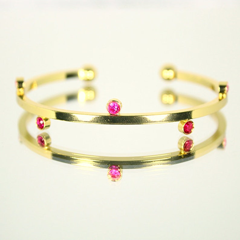 May include: A gold-colored bangle bracelet with multiple small, round, pink gemstones evenly spaced around the band. The bracelet is reflected in a shiny surface, creating a mirrored effect. The jewelry is simple and elegant.