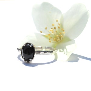 Puede incluir: Un anillo de plata con una piedra preciosa negra ovalada. El anillo tiene una banda texturizada y un diseño ajustable. Una flor blanca con estambres amarillos está en el fondo, sobre un fondo blanco brillante.