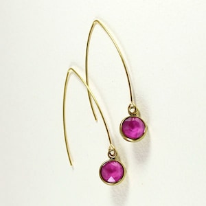 Peut inclure: Une paire de boucles d'oreilles pendantes dorées et incurvées. Chaque boucle d'oreille présente une pierre précieuse ronde de couleur magenta sertie dans une lunette dorée. Les boucles d'oreilles sont présentées sur fond blanc.