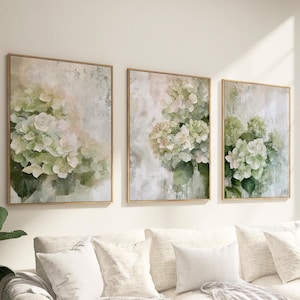 Op de afbeelding: Drie ingelijste wandkunstwerken met aquarel schilderijen van hortensia bloemen in groen, wit en roze tinten. Het kunstwerk heeft een textuur, abstracte achtergrond en is gevat in gouden lijsten. De schilderijen worden boven een crèmekleurige bank getoond.