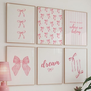 以下が含まれることがあります： ピンクと白の額縁に入った6枚のプリントのセット。リボンや弓、そして「dream」という言葉がデザインされています。プリントは白い壁に白いドレッサーの上に飾られています。