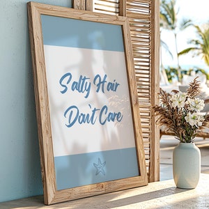 May include: A wooden framed print with the text "Salty Hair Don't Care" in blue script on a white background with a blue starfish at the bottom. The print is sitting on a wooden surface with a vase of white flowers in the background.