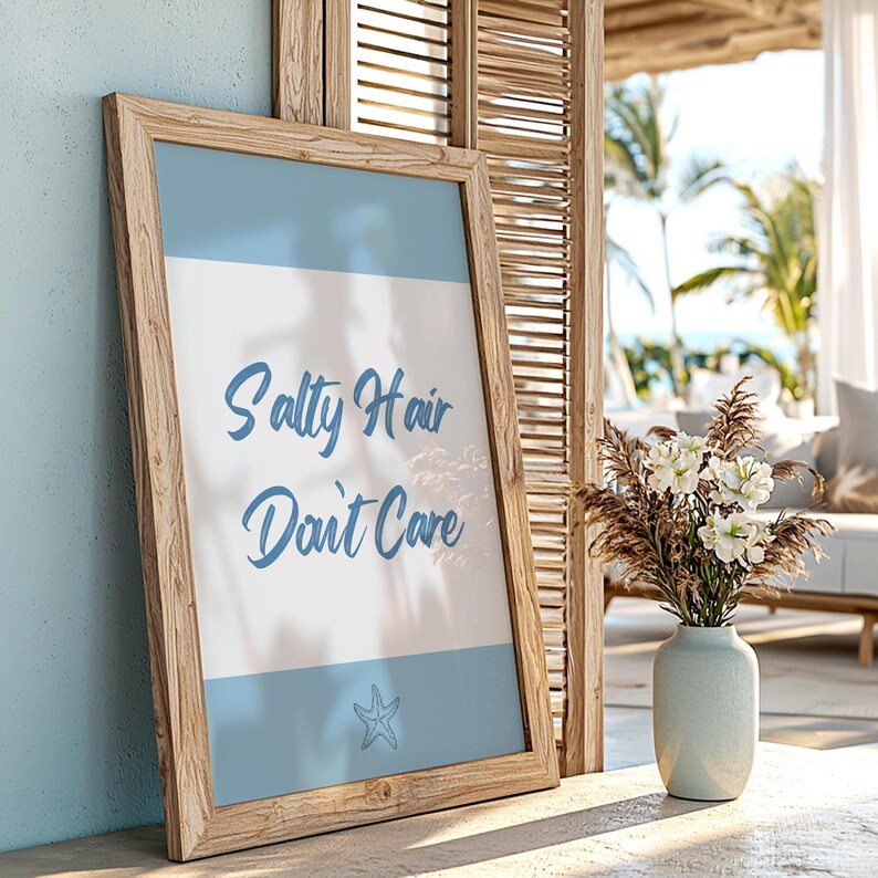 May include: A wooden framed print with the text "Salty Hair Don't Care" in blue script on a white background with a blue starfish at the bottom. The print is sitting on a wooden surface with a vase of white flowers in the background.