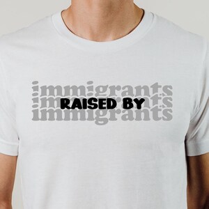 May include: White t-shirt with the text "Immigrants Raised By Immigrants" printed in black and gray.
