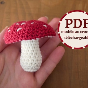 Puede incluir: Un hongo rojo y blanco tejido a crochet con un tallo blanco. La imagen incluye el texto "PDF modèle au crochet téléchargeable".