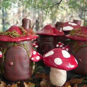 Peut inclure: Cinq maisons miniatures en forme de champignon avec des détails peints en rouge et blanc. Les maisons sont en argile et ont de la mousse et des vignes qui poussent dessus. Les maisons sont disposées en rangée sur un lit de feuilles d'automne.
