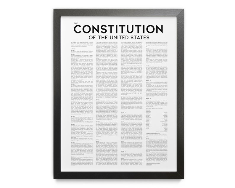 May include: A framed print of the United States Constitution. The black frame surrounds a white background filled with text. The title "THE CONSTITUTION OF THE UNITED STATES" is printed in bold black letters at the top.