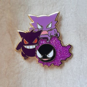 May include: A gold-colored enamel pin featuring three purple and black Pokemon characters: Gengar, Haunter, and a shiny, glittery Gastly.