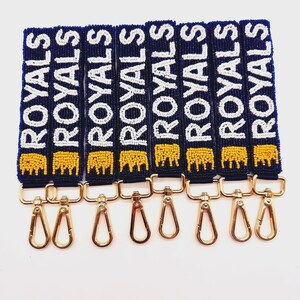 A collection of blue beaded straps with the word "ROYALS" in white beads. Each strap has a gold-colored clasp and a yellow beaded crow  at the bottom. The straps are displayed against a white background.