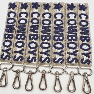 Beaded keychains/wristlets with the word "COWBOYS" in navy blue letters and a star. The keychains have silver-colored clasps. The background is a silver and white striped.