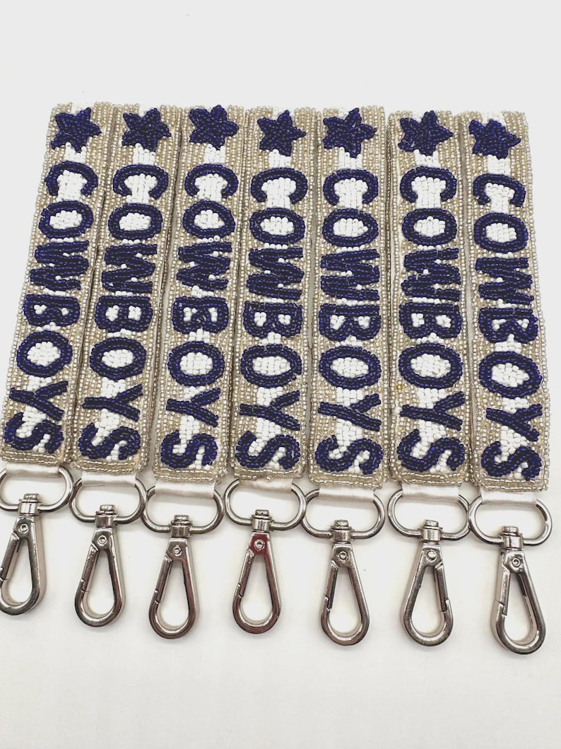 Beaded keychains/wristlets with the word "COWBOYS" in navy blue letters and a star. The keychains have silver-colored clasps. The background is a silver and white striped.