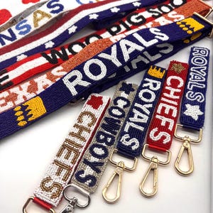 May include: A collection of beaded bag straps in various colors, featuring team names like "ROYALS" and "CHIEFS". The straps have gold-colored clasps and are decorated with stars and hearts.