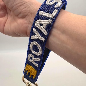 May include: A wristlet keychain featuring a blue beaded strap with the word ROYALS spelled out in white beads and a yellow beaded crown icon at the base. The strap is attached to a gold-toned metal lobster clasp hardware for secure attachment.