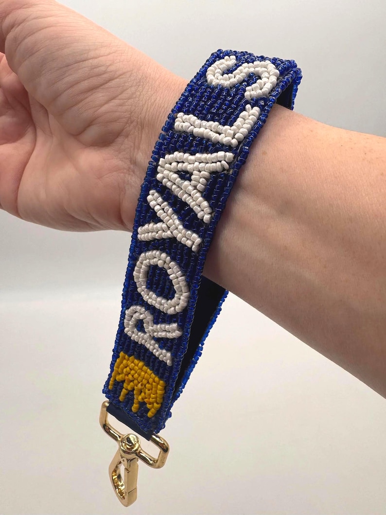 May include: A wristlet keychain featuring a blue beaded strap with the word ROYALS spelled out in white beads and a yellow beaded crown icon at the base. The strap is attached to a gold-toned metal lobster clasp hardware for secure attachment.
