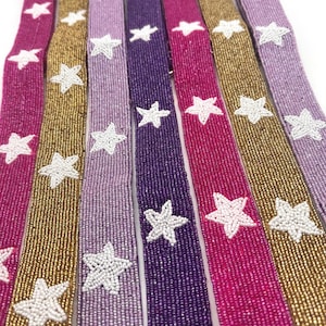 May include: A collection of beaded belts in various colors, including gold, purple, and pink. Each belt is adorned with white star-shaped embellishments. The belts are arranged side by side, showcasing the range of colors and designs.