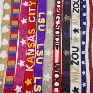 May include: A collection of ten beaded straps featuring various designs including white hearts on gold, rainbow stripes, white stars on pink, the text KANSAS CITY, the text LSU, white stars on silver, the text WOO PIG SOOIE, purple stars on black, the text MIZZOU, and a red, white, and blue star pattern.
