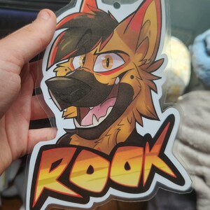 May include: A clear plastic badge featuring a colourful illustration of a cartoon wolf with red, yellow and black fur. The wolf is smiling and has a black nose. The text "ROOK" is printed in yellow, orange and red letters on the badge.