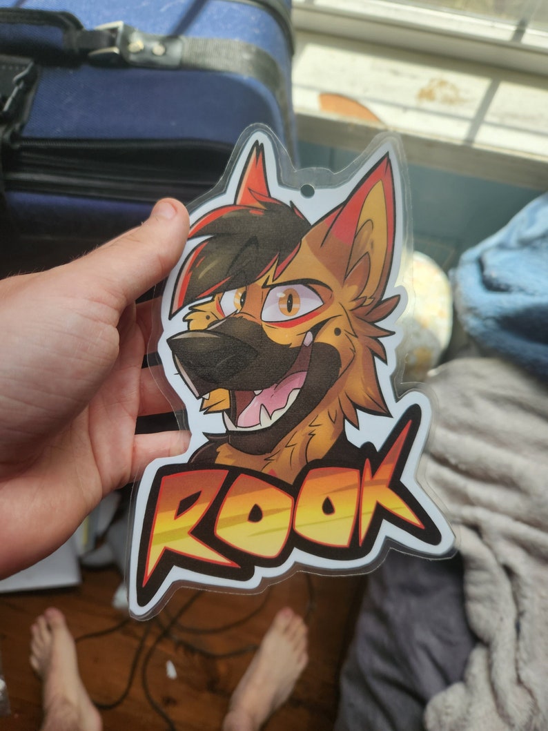 May include: A clear plastic badge featuring a colourful illustration of a cartoon wolf with red, yellow and black fur. The wolf is smiling and has a black nose. The text "ROOK" is printed in yellow, orange and red letters on the badge.