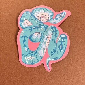 May include: A sticker featuring a stylized illustration of a snake in shades of teal, pink, and white. The snake is coiled and has a detailed pattern. The sticker has a pink border and is set against a brown background.