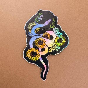 May include: A holographic sticker featuring a stylized snake intertwined with sunflowers and other floral elements. The snake is primarily blue and pink, with yellow sunflowers and green foliage. The sticker has a black border and is set against a brown background.