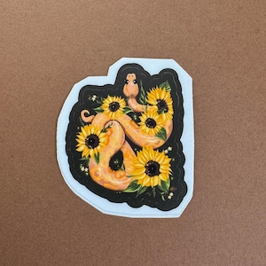 May include: A sticker featuring a stylized illustration of a snake intertwined with sunflowers. The snake is a warm orange color, and the sunflowers have yellow petals and dark centers. The sticker has a black border and a white outline.