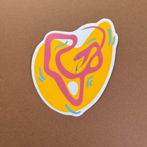 May include: A die-cut sticker featuring a stylized snake design. The snake is pink and curves across a yellow background. Blue accents and a white border complete the design, set against a brown backdrop.