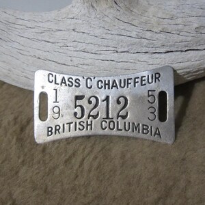 Könnte beinhalten: Ein silbernes Metallschild mit dem Text "CLASS 'C' CHAUFFEUR" und der Nummer "5212" darauf gedruckt. Das Schild trägt auch den Text "BRITISH COLUMBIA" aufgedruckt.