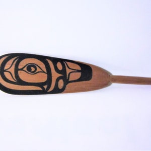 May include: A wooden paddle with a black and white design of a stylized face. The paddle is about 18 inches long and has a rounded end.