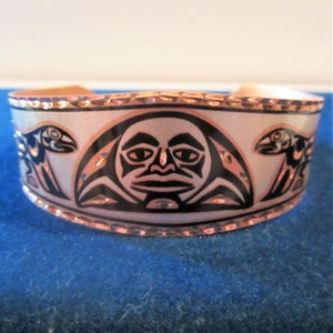 May include: A copper bracelet with a white background and black design. The design features a stylised face with two birds on either side.