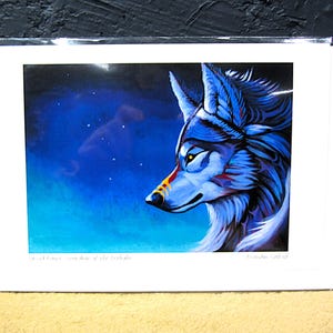 May include: A print of a blue wolf with yellow eyes and red accents, set against a dark blue and starry background. The artwork is framed in a white border. Text at the bottom reads "Sacred Spirit - Guardian of the Twilight" and the artist's name.