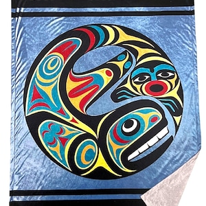 May include: A blue and black fleece blanket with a circular design featuring a stylised salmon in a traditional Northwest Coast Indigenous art style. The salmon is depicted in red, yellow, blue, and green colours.