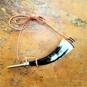 Könnte beinhalten: Ein schwarzes und weißes Hornpuderhorn mit einem Messingspitz und einem Lederriemen. Das Horn befindet sich auf einer Holzoberfläche.