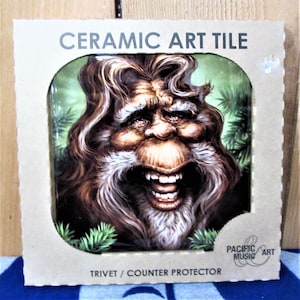 May include: A ceramic art tile featuring a cartoon-like illustration of a smiling, bearded man with long hair. The tile is packaged in a cardboard box with the text "Ceramic Art Tile" and "Trivet/Counter Protector" printed on it. The box also has a logo for "Pacific Art Music".