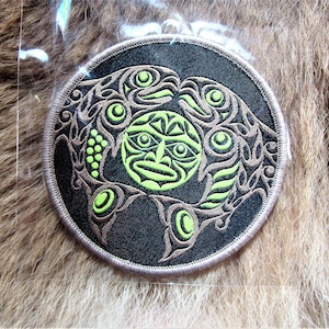 Può includere: Un cerotto rotondo in tessuto nero con un disegno ricamato verde e marrone di un viso stilizzato circondato da due pesci. Il design ricorda l'arte dei nativi americani.