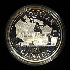 May include: A silver Canadian dollar coin from 1981. The coin features a detailed image of a steam train on tracks, with the word "DOLLAR" at the top and "CANADA" at the bottom. The outline of Canada is in the background.