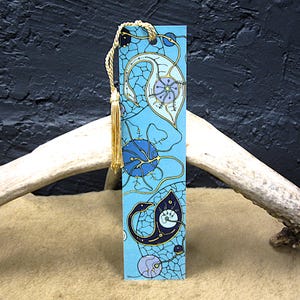 May include: A blue bookmark with an abstract design featuring organic shapes in blue, purple, and gold. The bookmark has a gold tassel attached to the top. The background is a dark blue.