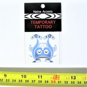 Könnte beinhalten: Ein temporäres Tattoo mit einem stilisierten Design in Blau und Weiß, das eine Kreatur mit großen Augen und einem breiten Mund darstellt. Das Tattoo ist in einer weißen Karte verpackt, auf der der Text "Native Accents" und "Temporary Tattoo" auf schwarzem Hintergrund gedruckt ist.