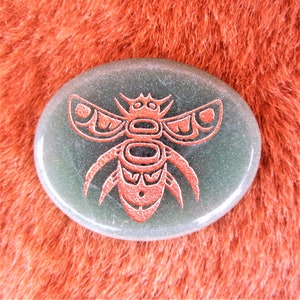 May include: A green oval-shaped stone with a copper-coloured stylised bee design. The bee has a geometric pattern and is centred on the stone.