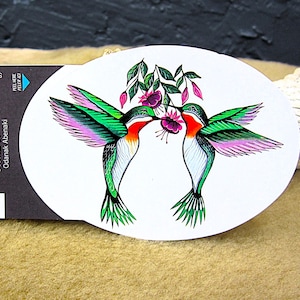 May include: Oval sticker featuring two hummingbirds with green wings, pink accents, and red throats, kissing near pink flowers. The sticker has the text "Sweet Duo" and the names "Jessica Somers" and "Odanak Abenaki".