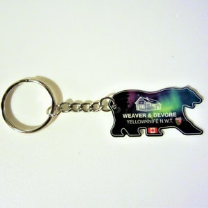 May include: A silver bear-shaped bottle opener keychain with a colorful aurora borealis design. The keychain features the text "WEAVER & DEVORE YELLOWKNIFE N.W.T." and a Canadian flag.