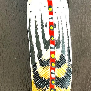 May include: A hand-painted feather with black, white, red, and yellow details. The feather is painted with a design of a stylized bird or animal.