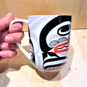 May include: White ceramic mug with a black, red and grey design featuring a stylised face. The design is reminiscent of Northwest Coast Indigenous art.