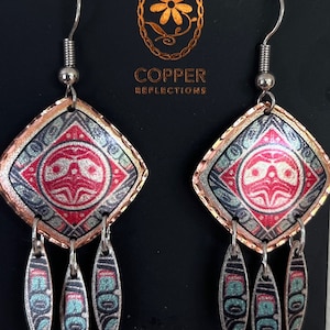 May include: Copper earrings with a geometric design featuring a red and white stylized face with a copper border. The earrings have three silver teardrop-shaped charms hanging from the bottom.