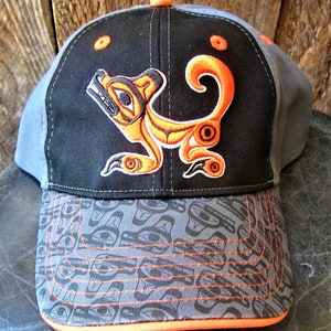 May include: A black and gray baseball cap with an orange and white embroidered design of a stylized animal. The cap has a gray and black patterned brim.