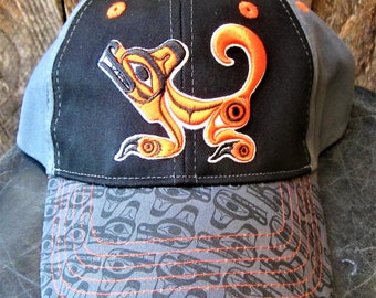 May include: A black and gray baseball cap with an orange and white embroidered design of a stylized animal. The cap has a gray and black patterned brim.