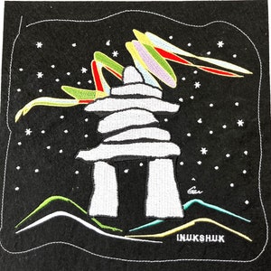 Può includere: Una toppa ricamata in feltro bianco che raffigura un inukshuk, una struttura in pietra tradizionale Inuit, sotto un'aurora boreale colorata su uno sfondo nero. Il testo "INUKSHUK" è ricamato in bianco nella parte inferiore della toppa.