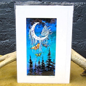Op de afbeelding: Een ingelijste kunstdruk met een halve maan met een dromenvangerontwerp en verschillende kleurrijke vlinders. Het kunstwerk is geplaatst tegen een blauwe en blauwgroene achtergrond met silhouetbomen. De print is gesigneerd door de kunstenaar.