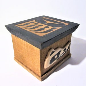 May include: A wooden box with a black and tan geometric design on the top and a black and white animal design on the side. The box is made of wood and has a dark gray stain.