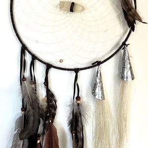 May include: A white dreamcatcher with brown leather straps and feathers. The dreamcatcher has a crystal in the center and is decorated with silver bells.